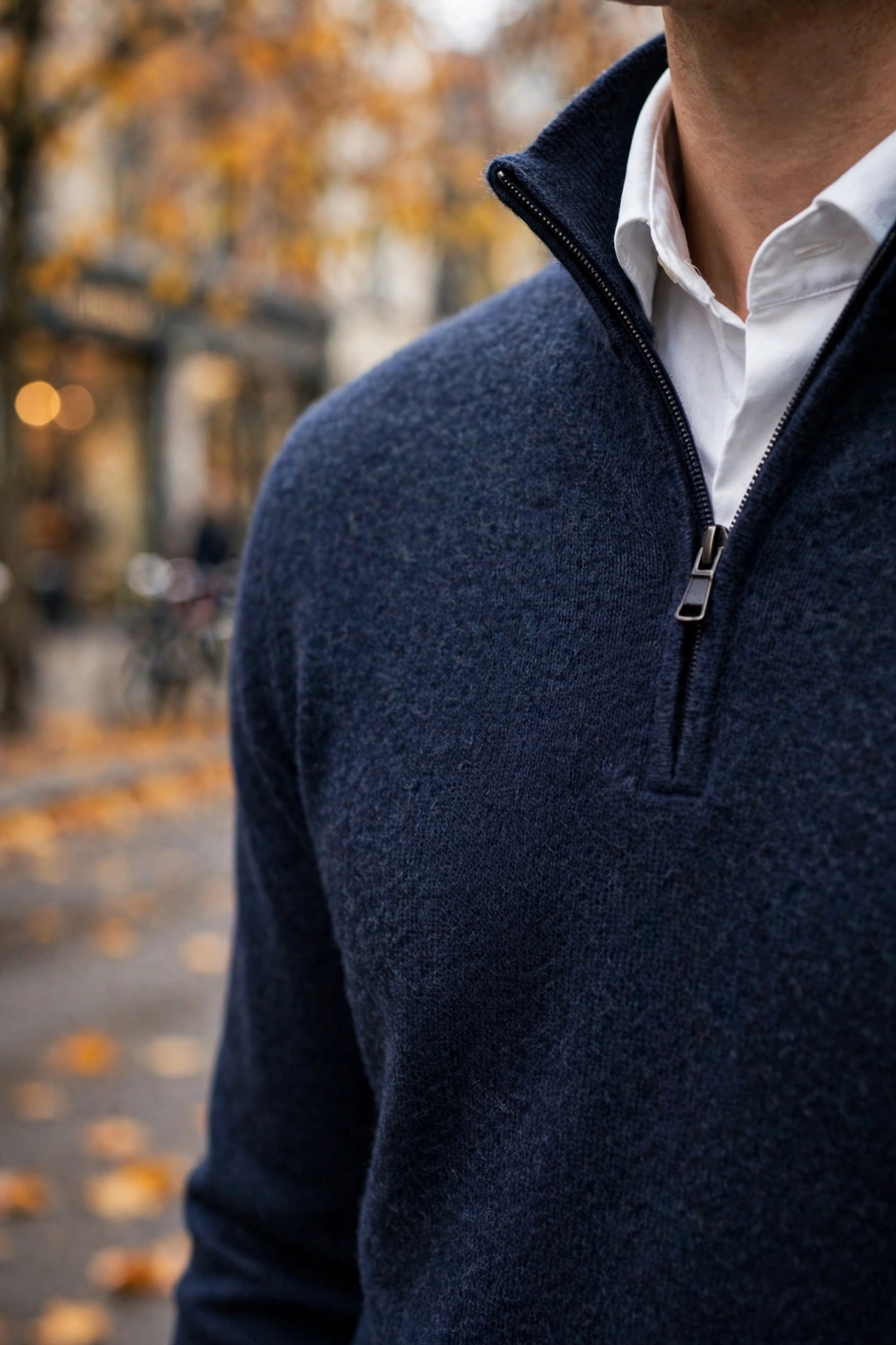 Close-up of a man wearing a navy merino quarter-zip sweater over a white collared shirt on an autumn city street